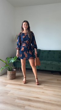 Kratka obleka s kratkimi rokavi v mornarski modri barvi z abstraktnimi rjavimi vzorci, zategnjenim pasom in razširjenimi rokavi. Dodatki vključujejo rjavo torbico in krema salonarje.