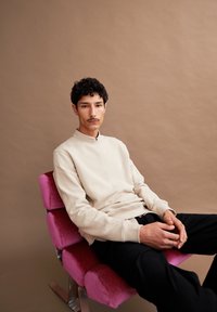 Beige Crewneck-Sweatshirt mit minimalistischem Design, auf einem pinken Samtsessel sitzend. Schwarze Hosen, sichtbares Tattoo an den Händen. Neutraler Hintergrund.