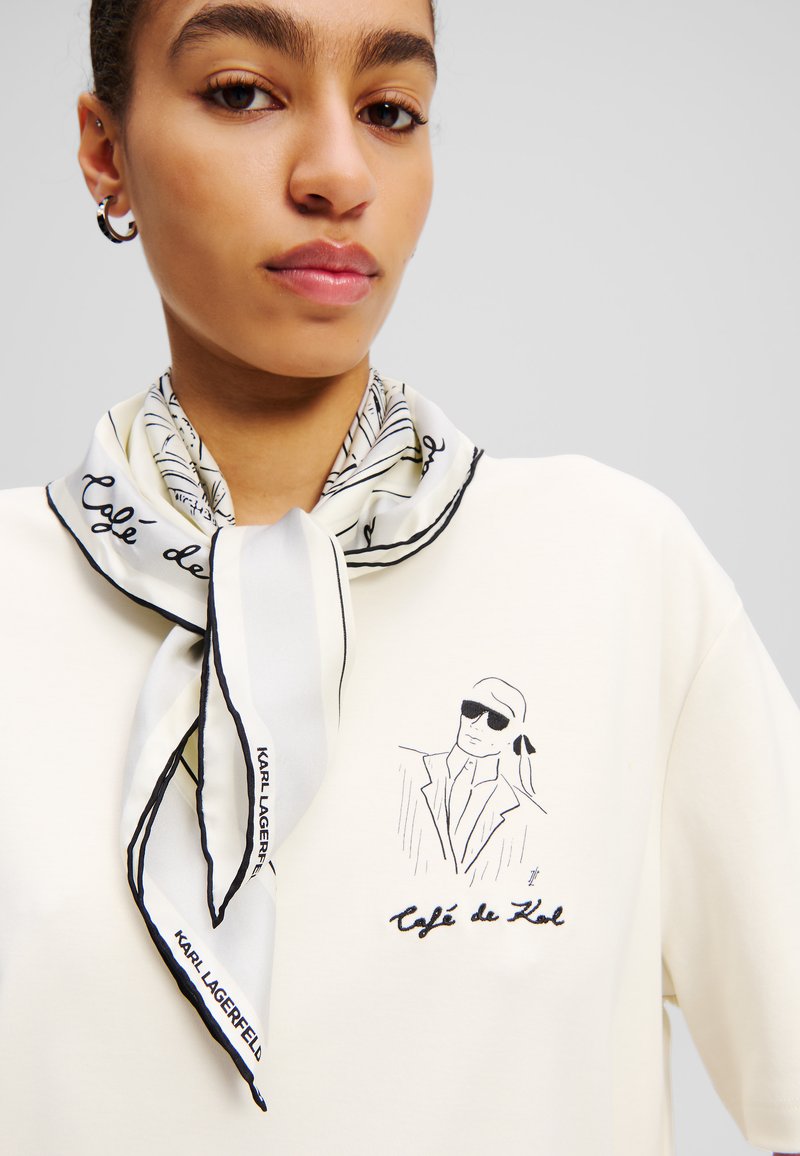 Fehér pamut pulóver, amelyen egy napszemüveget és dzsekit viselő figura van nyomtatva, kiegészítve egy fekete szegélyű mintás selyemsállal.