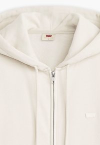 Kreem värvi lukuga kapuutsiga sviiter, mis on valmistatud pehmest kangast. Omab vooderdatud kapuutsi, nööre ja Levi's märgist. Sisaldab metallset lukku, millel on brändingud.