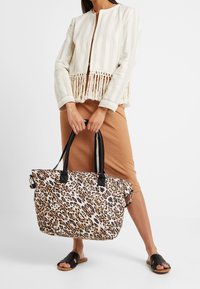 Sac fourre-tout en tissu imprimé animal avec des poignées noires, présentant un motif léopard audacieux. Le modèle porte une veste crème rayée à franges et une jupe marron.