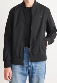 Schwarze Bomberjacke aus leichtem Stoff, mit Frontreißverschluss, gerippten Bündchen und Seitentaschen. Wird über einem schwarzen Oberteil und blauen Jeans getragen.