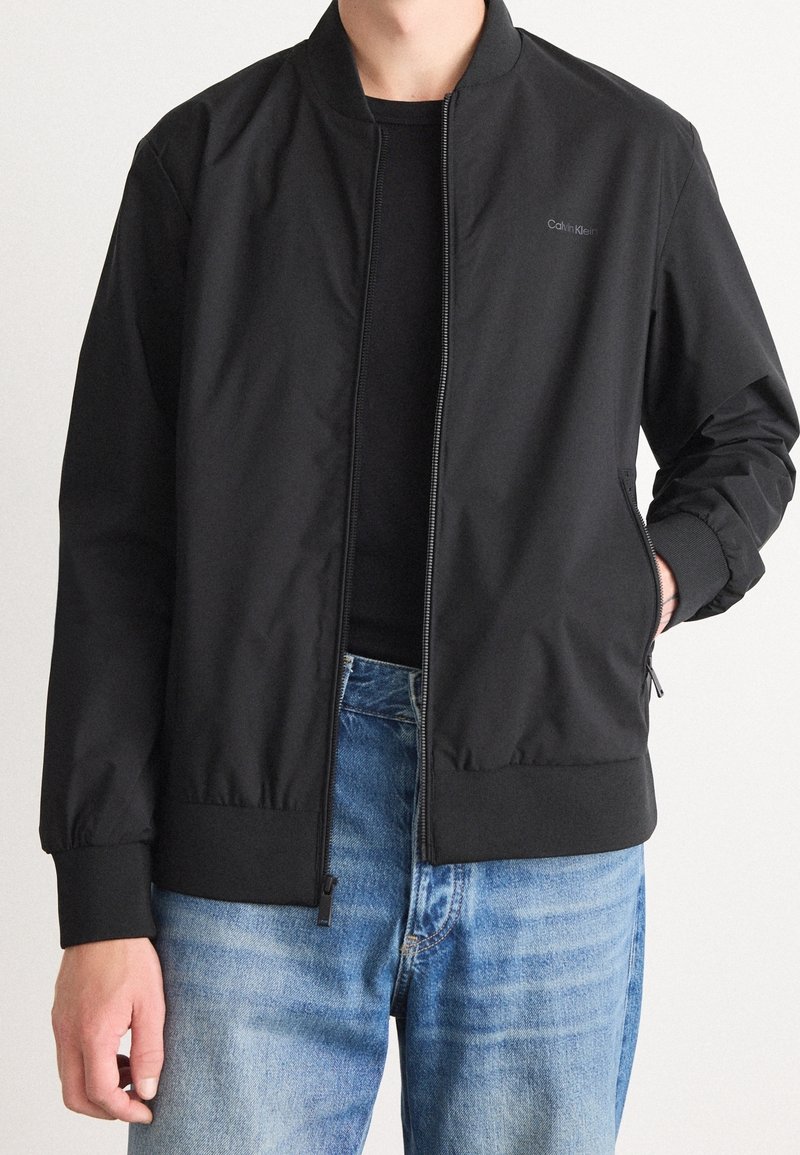 Schwarze Bomberjacke aus leichtem Stoff, mit Frontreißverschluss, gerippten Bündchen und Seitentaschen. Wird über einem schwarzen Oberteil und blauen Jeans getragen.