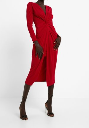 Czerwona sukienka z długim rękawem, dekoltem w kształcie litery V i zwiniętym przednim detalem, o dopasowanym kroju i długości do kolan. W zestawie z czarnymi butami na wysokim obcasie.