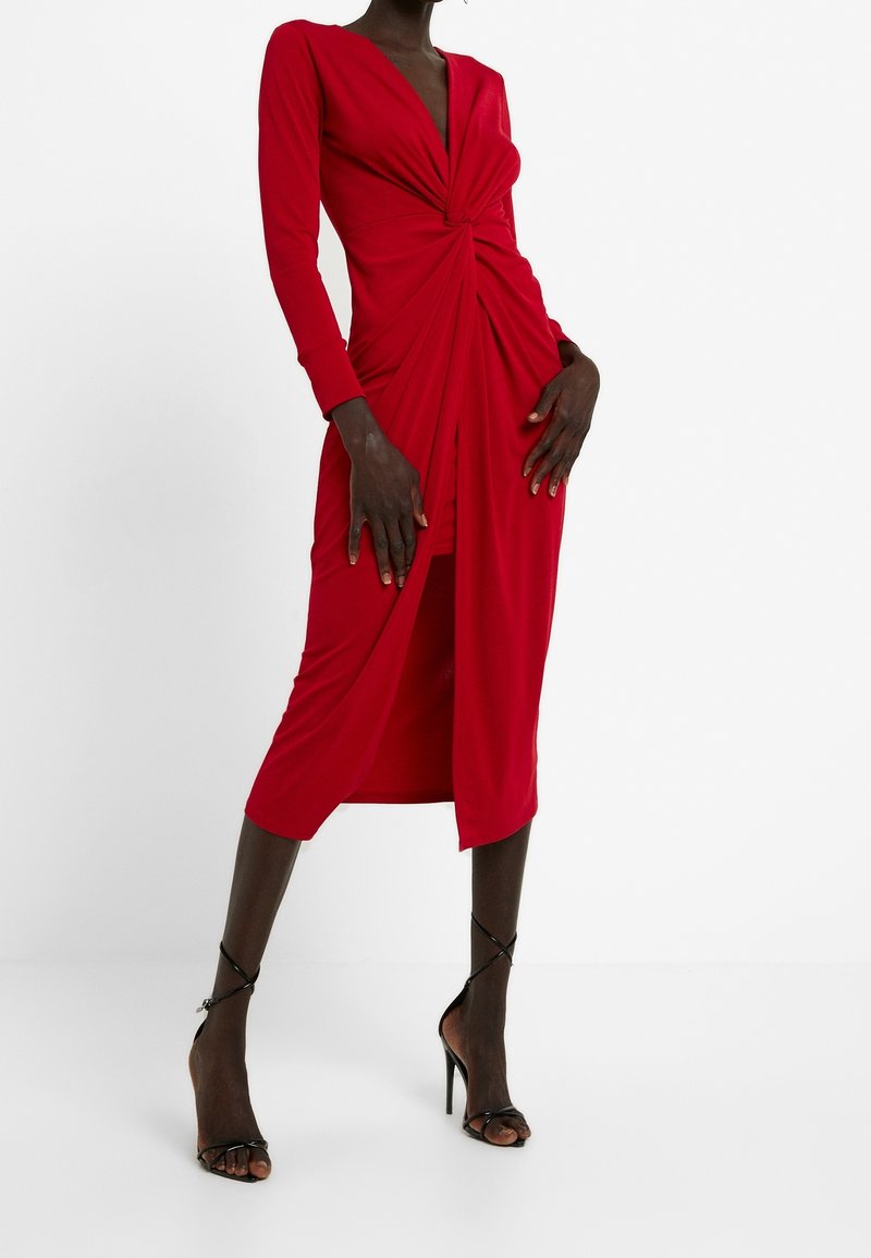 Robe rouge à manches longues avec un col en V et un détail torsadé à l'avant, présentant une silhouette ajustée et un ourlet arrivant au genou. Associée à des chaussures noires à talons hauts.