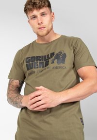 Alyvuogių spalvos medvilninis marškinėliai su ryškiu juodu grafiniu spaudiniu, kuriame užrašyta "GORILLA WEAR" ir "JAV", trumpomis rankovėmis, apvaliu kaklu.