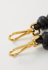 Sección de cuero negro con textura anudada, que presenta clips de metal dorado y herrajes detallados con el nombre de marca en relieve "Coccinelle."