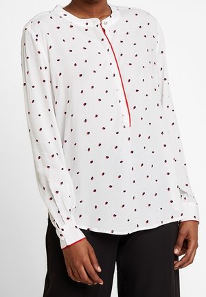 Blouse blanche à manches longues avec petits pois rouges et noirs, bordure rouge le long de la patte de boutonnage et aux poignets, portée avec un pantalon noir.