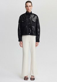 Jachetă scurtă din paiete negre, cu închidere cu butoane și două buzunare pe piept, asociată cu pantaloni albi evazați și pantofi cu toc subțire negri.