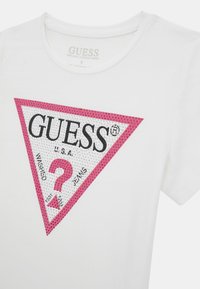 T-shirt en coton biologique blanc avec un logo triangle rose portant les inscriptions "GUESS", "U.S.A" et un point d'interrogation en noir sur un fond texturé.