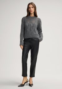 Woman wearing a grey knit sweater and black leather pants with black high-heeled slingback shoes, standing with one hand in pocket.