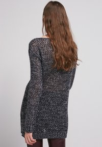 Lång, löst stickad tröja i mörkgrått med ljusa och mörka prickar. Har en bred halsringning och ribbad nederkant. Designad för en avslappnad passform.