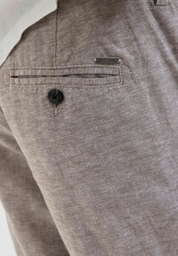 Smeđe lanene hlače sa stražnjim džepom ukrašenim crnim dugmetom i srebrnom oznakom "JACK & JONES". Teksturirani materijal s blagim sjajem.