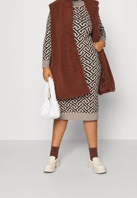 Gilet sans manches marron sur une robe tricotée à motif géométrique ; sac en fausse fourrure blanche ; mocassins épais crème ; chaussettes marron.
