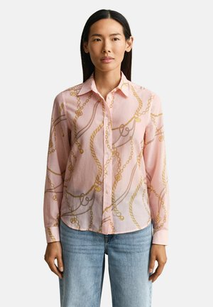 Frau trägt ein hellrosa Hemd mit Knöpfen und einem goldenen Kettenmuster sowie hellblaue Jeans, steht vor einem schlichten weißen Hintergrund.