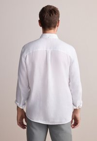 Chemise blanche en lin à manches longues, avec des poignets boutonnés, détail de yoke au dos et texture subtile, portée avec un pantalon gris.