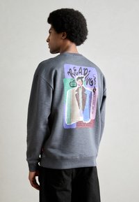 Sivi dukserica s okruglim izrezom, koja ima šareni grafički prikaz na leđima, s tekstom i uzorcima u raznim nijansama.