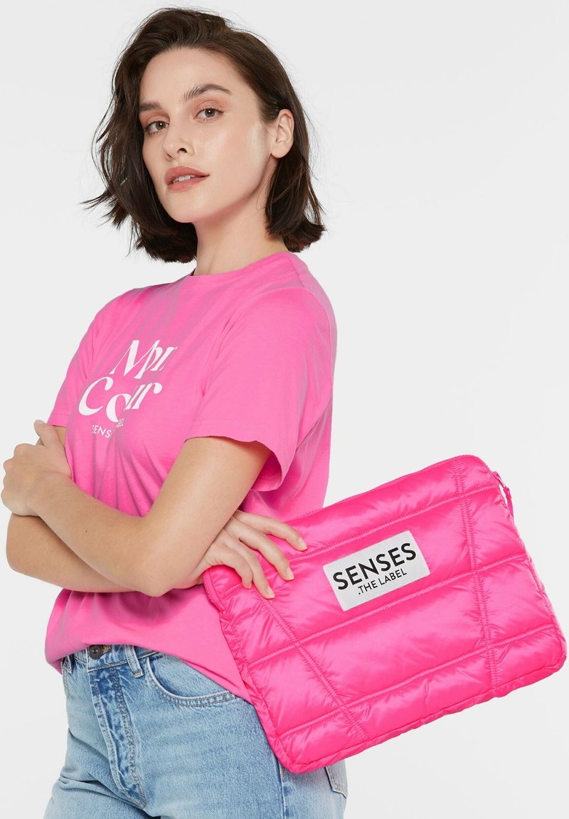 Helle pinke gepolsterte Laptop-Tasche mit glatter Oberfläche, die ein auffälliges Etikett mit der Aufschrift "SENSES THE LABEL" auf der Vorderseite trägt.