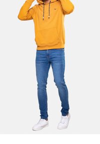 Felpa gialla brillante con lacci, tasca frontale; abbinata a jeans blu aderenti e sneakers bianche. Design semplice, vestibilità casual.