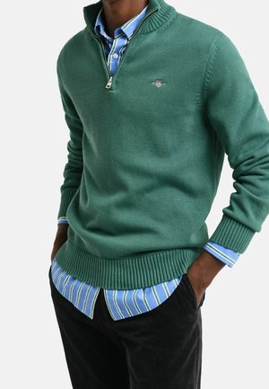 Homme portant un pull vert à fermeture éclair quart de longueur sur une chemise rayée bleu et blanc à boutons, avec un pantalon noir, mains dans les poches.
