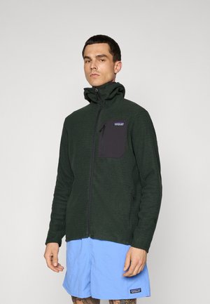 Man som bär en mörkgrön zip-up fleecejacka med hög krage och ljusblå shorts, stående mot en enkel ljus bakgrund.