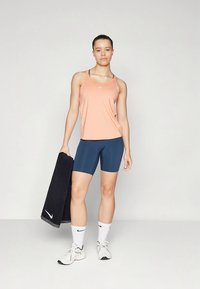 Top desportivo rosa, calções de bicicleta azul-marinho, sapatos desportivos brancos, segurando uma toalha preta. Apresenta o logótipo da Nike no top e nos sapatos.