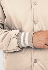 Beżowa kurtka bombowca o gładkim wykończeniu. Posiada prążkowane mankiety z trzema poziomymi paskami w bieli i szarości. Zawiera pasujące guziki.