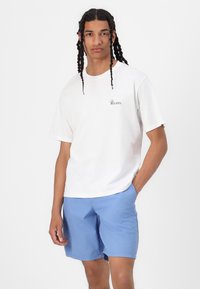 Camiseta blanca de algodón con mangas cortas, que presenta un pequeño texto gráfico en negro. Combinada con pantalones cortos azul claro, de tejido suave y ajuste casual.