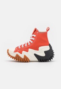 Sneaker alta in tela rossa con lacci bianchi, suola testurizzata alternante arancione e nera, design a zigzag e linguetta sul tallone.