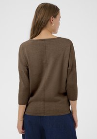 Brauner Pullover mit weitem Ausschnitt und Dreiviertelärmeln. Er hat eine gerippte Textur und einen geraden Schnitt, kombiniert mit blauen Jeans.