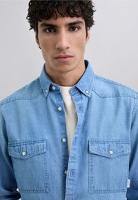 Camisa de denim azul claro con cuello abotonado, dos bolsillos frontales y botones blancos. La tela parece suave con una textura lisa.