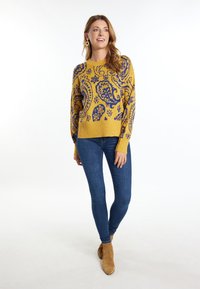 Mosterdgele gebreide trui met een blauw paisleypatroon, gecombineerd met donkerblauwe skinny jeans en tan enkellaarzen.
