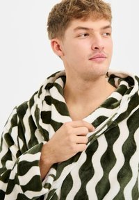 Grøn- og hvidbølget badekåbe i blødt plysmateriale med hætte, vide ærmer og frontåbning med en synlig hånd.