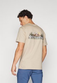 Cămașă t-shirt din bumbac bej cu un grafic pe spate care reprezintă un peisaj montan, cu copaci verzi, munți gri și accente solare.