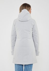 Donna che indossa un cappotto invernale grigio chiaro con cappuccio e jeans blu, in piedi con la schiena rivolta verso la fotocamera su uno sfondo semplice.
