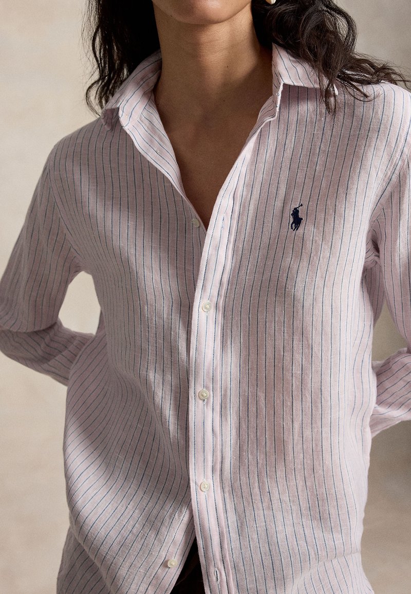 Femme portant une chemise à manches longues de couleur rose clair avec de fines rayures verticales bleues et un logo brodé sombre d'un joueur de polo sur la poitrine.