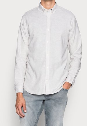 Mężczyzna w białej, zapinanej na guziki koszuli z długim rękawem i jasnoniebieskich dżinsach, z jedną ręką w kieszeni, a drugą swobodnie opuszczoną wzdłuż ciała.