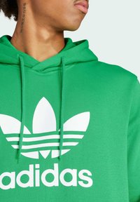 Sudadera verde de algodón, con un gran logo blanco de Adidas que presenta tres rayas y acentos de diseño floral. Detalle de cordones presente.