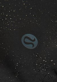 Nærbillede af vanddråber, der perler på sort vandtæt stof med et cirkulært, stiliseret "S"-logo i midten.