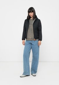 Zwarte gewatteerde jas met capuchon, voorzien van ribbelstructuur en oranje accent, gedragen over een grijze sweatshirt. Gecombineerd met ruime blauwe jeans.