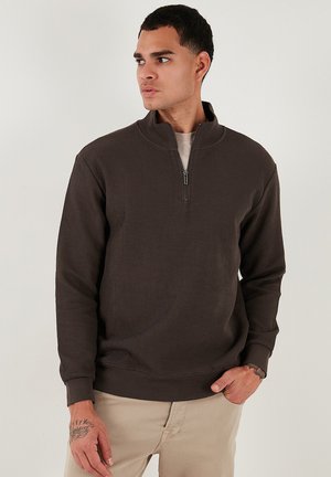 Sudadera de media cremallera de color marrón oscuro, con textura acanalada, mangas largas, corte relajado, llevada sobre una camisa de color claro, con aberturas laterales en el dobladillo.