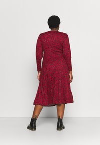 Robe rouge à manches longues avec un corsage ajusté et un ourlet évasé, présentant un motif abstrait noir et bleu. Associée à des bottines noires.