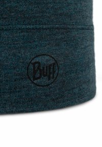 Close-up of dark teal knitted fabric with black "Buff" logo printed inside a circle near the seam edge on a winter accessory.