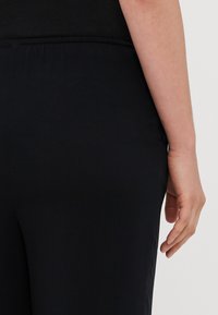 Sorte tights med jevn tekstur, med elastisk midje og en tettsittende silhuett, vist fra bak, med hånden på hoften.