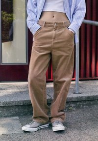 Pantalons en velours côtelé marron à coupe ample, avec une fermeture à bouton, des poches arrière et des côtes verticales subtiles. Associés à des baskets grises.