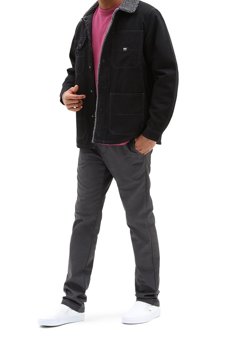 Homme portant une veste noire doublée sherpa, une chemise rose, un pantalon gris et des chaussures blanches sans lacets, debout avec une main dans la poche.