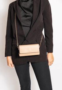 Beige schoudertas met een gladde textuur, rechthoekige vorm, magnetische klepsluiting en kettingband, gedragen over een zwarte blazer.