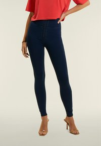 Leggings de tiro alto en color navy, confeccionados en tejido elástico, con un cierre frontal y detalles en los tobillos, combinados con una blusa roja suelta y tacones abiertos.