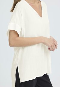 Top bianco a maniche corte con scollatura a V, caratterizzato da una texture a coste, spacchi laterali e una silhouette rilassata, abbinato a pantaloni neri.
