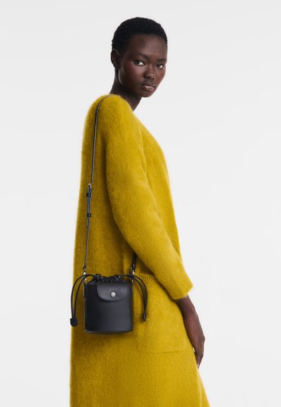 Schwarze Leder-Eimer-Tasche mit Zugverschluss und silberner Hardware. Glatte Textur, kompaktes Design, getragen zu einem leuchtend gelben Strickpullover.